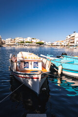 A small wooden boat with a striped canopy and a turquoise boat docked near a waterfront town with whitewashed buildings and outdoor cafes in Crete. © Aerial Film Studio
