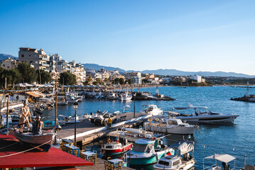A harbor on Crete, Greece, features small boats, yachts, pastel buildings, lush greenery, distant mountains, and a fisherman statue under a clear sky.