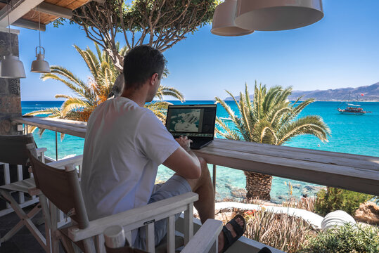 A man sits on a wooden chair with a laptop on a shaded balcony. The view includes turquoise sea, a distant boat, palm trees, and a clear blue sky.