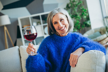 Charming senior woman in a blue sweater enjoying wine on a cozy couch in a bright modern living room setting