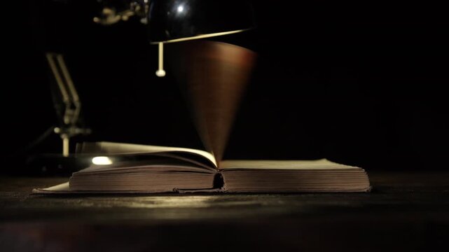 Pages of old book turning under light of table lamp on black background close-up