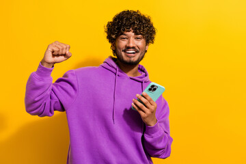 Naklejka premium Cheerful young man in a purple hoodie celebrates while holding a smartphone, against a vibrant yellow background