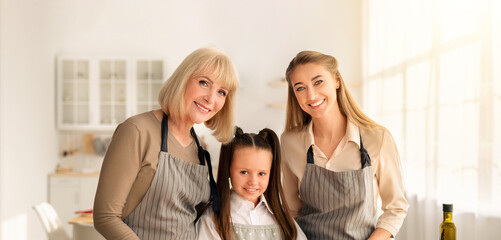 Portrait of multigenerational family with cute girl, her mom and granny posing in kitchen, smiling at camera. Young woman with her daughter and elderly mother cooking dinner at home