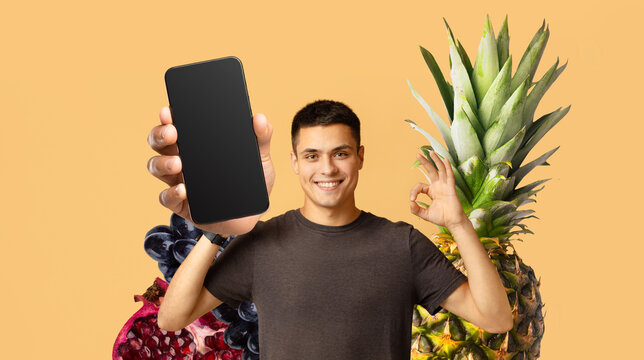 A young man smiles and holds up a smartphone in front of colorful fruits. The vibrant collage shows his enthusiasm for healthy eating and travel experiences. The setting is bright and cheerful.