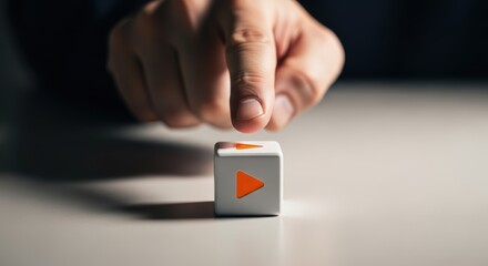 A finger hovering over a white cube with an orange play button on it on a white surface in soft light