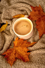 A white mug of hot coffee sits in a knitted blanket with yellow autumn leaves.