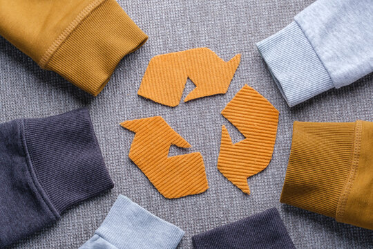 Sweater sleeves in shades of gray, mustard, and light blue surround a recycling symbol made from orange fabric. Concept of clothing recycling, sustainable fashion, and textile waste reduction.