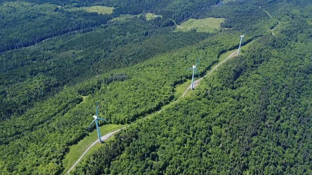 A view of three operating wind turbines connected by a forest trail atop a remote, verdant mountain. Saint-Leandre, Quebec, Canada, 2025.