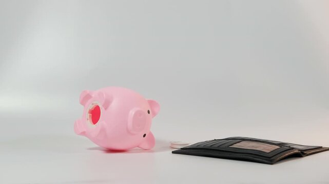 A man is emptying his wallet onto a white surface. The scattered coins and dollar bill surround an empty pink piggy bank, indicating savings efforts, slow motion