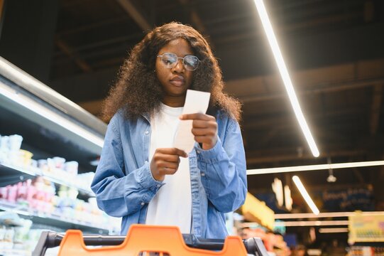 african american woman in a grocery store holding a receipt and checking the prices of products. The concept of rising prices for products - Powered by Adobe