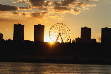 Sunset view behind a ferris wheel and city skyline. Orange sky with clouds.