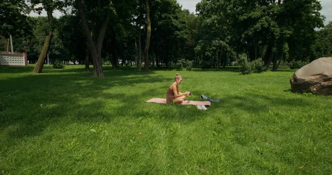 A woman turns on a video tutorial on her tablet and starts Pilates sitting on a mat in the park.