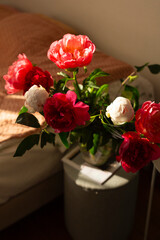An enchanting mix of pink, white, and red peonies in a glass vase, artfully arranged on a small table, illuminated by the rich, glowing hues of sunset.