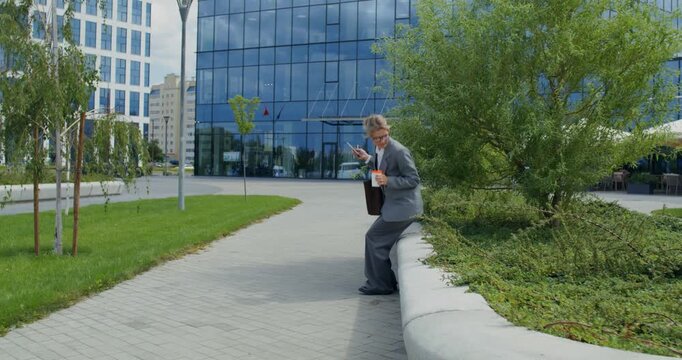 A woman sits on a parapet on a city street and uses a mobile phone. A businesswoman uses a mobile phone near an office building.