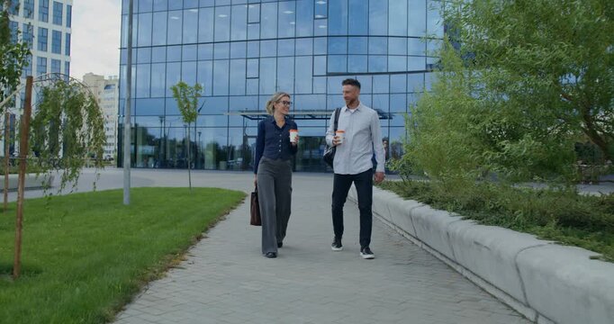 A man and a woman drink coffee from disposable cups and talk sitting on the parapet near an office building.