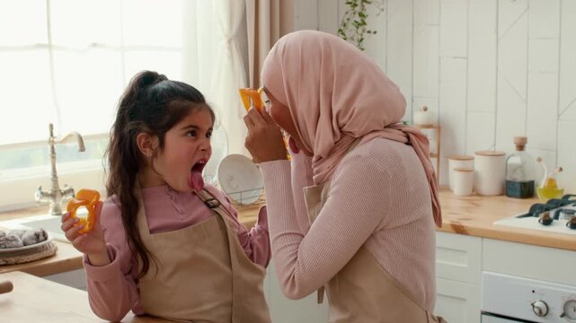 A mother and daughter share joyful and affectionate moments in a bright kitchen, playing with colorful vegetables while wearing matching aprons. Their laughter fills the warm atmosphere.