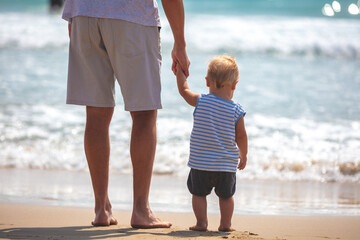First steps on the beach. A little boy and his father are walking on the beach on a sunny summer day. The father is holding his son's hand. Rear view