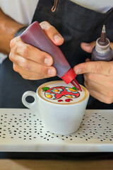 Barista creating vivid latte art with heart using colored syrups on coffee foam in white cup. Concept of latte art, barista skill and coffee design creativity