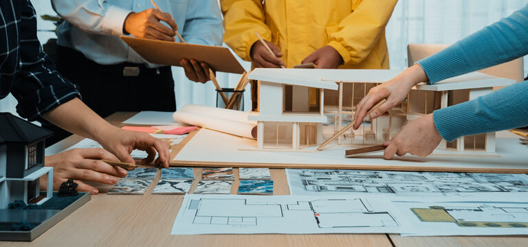 A diverse team of professionals engages in an architectural design meeting, reviewing blueprints and 3D models in a bright, modern workspace. SACTR - Powered by Adobe