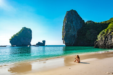 Woman in a swimsuit sitting on the sand of Nui Beach in Krabi, Thailand, enjoying the serene ocean view with limestone karsts in the background and longtail boats floating on the turquoise water