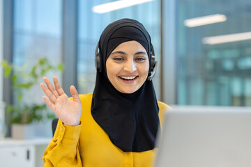 Muslim businesswoman wearing a headset waving hand during an online video call, providing remote customer service or participating in virtual meeting from modern office