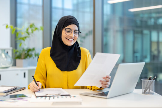 Smiling muslim businesswoman wearing a hijab and glasses, reviewing financial data on paper documents and making notes while working diligently on a laptop in a modern office