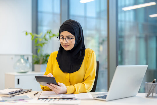 Muslim businesswoman wearing a black hijab and glasses, working in a modern office, concentrating while using a calculator for financial planning and accounting tasks on her desk