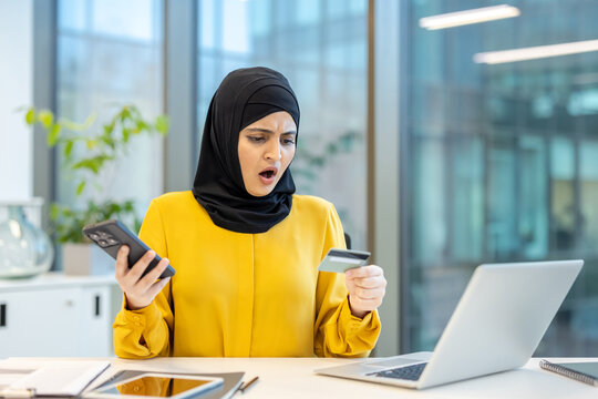 Arabian woman wearing a hijab sitting at an office desk, holding a credit card and smartphone, showing a shocked expression due to an online shopping payment problem or fraud - Powered by Adobe