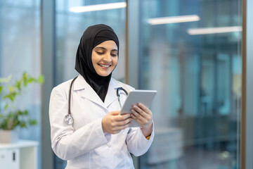 Young arabian female doctor in hijab and white coat, smiling while using a tablet to review electronic health records and manage patient data in a modern clinic setting
