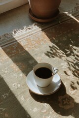 A cup of black coffee rests on a vintage patterned rug, illuminated by warm morning sunlight streaming through a nearby window. The soft shadows of leaves add a calm, poetic atmosphere