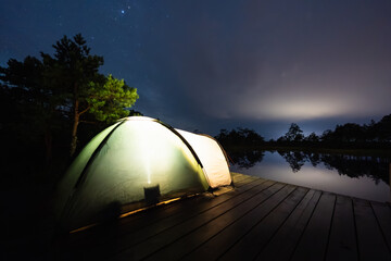 Illuminated camping tent on a wooden platform by a lake in an Estonian bog under a starry night sky.