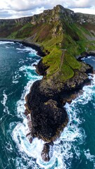 High-angle view of dramatic coastal headland, rugged cliffs, and powerful waves. Lush green vegetation crowns the top of a dark basalt rock promontory jutting into a choppy, dark-blue ocean