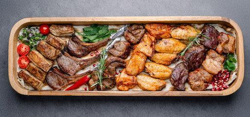 Assorted grilled meat platter with vegetables. Top view of assorted grilled meat cuts served on a wooden tray with vegetables, herbs, and spices.