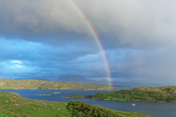 Double rainbow over the islands in Badcall Bay with dark clouds and the Quinag mountain range in shadow in the background.  Sheep and goats are grazing on the grass in the foreground