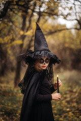 A witch girl with skull makeup holds a candle in a dark forest on Halloween