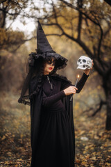 A witch girl with skull makeup holds a candle in a dark forest on Halloween