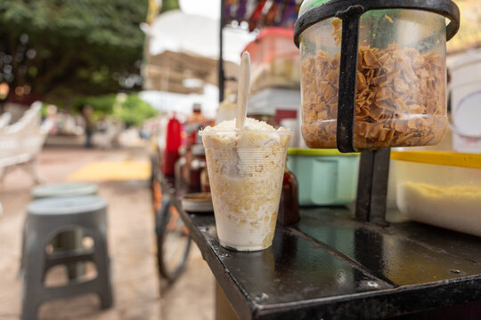 Traditional Mexican esquite in a cup, with cooked corn kernels, cream, and cheese, at a street stall in a garden. chascas, vasolotes, trolelotes, elote en vasito, elote en vaso