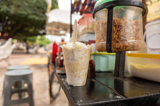Traditional Mexican esquite in a cup, with cooked corn kernels, cream, and cheese, at a street stall in a garden. chascas, vasolotes, trolelotes, elote en vasito, elote en vaso