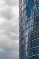 Obraz premium Modern glass skyscraper facade with blue reflective windows against a cloudy sky. Contemporary office building architecture with curved glass surface reflecting clouds
