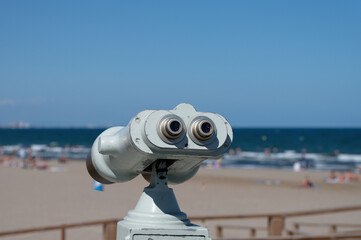 A coin-operated binoculars showing the sandy Playa de las Arenas beach in Valencia, Spain on a sunny day with beachgoers and the sea