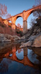 A stone arch bridge reflected serenely in a tranquil river at sunset, showcasing intricate architecture and a serene autumnal ambiance.