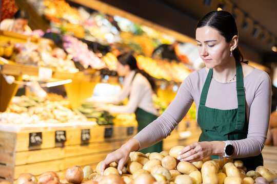 Female seller carefully lays out seasonal potatoes on the counter of the vegetable section in the supermarket. Grocery store worker is standing in the department with vegetables and checking potatoes