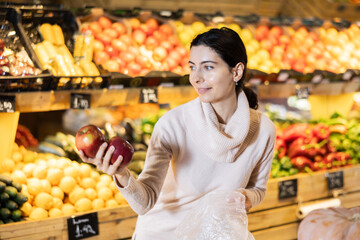 Near shelves and display cases in sales area of store, woman customer takes ripe apple out of box. Wide range of agricultural products. Self-service, comfortable shopping.