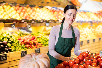 Positive middle-aged female seller putting tomatoes on stand in fruit and vegetable market
