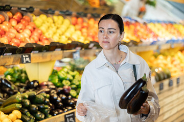 Attentive middle-aged female buyer holding eggplants in large fruit and vegetable market