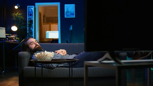 Smiling man lounged on couch at home eating popcorn while browsing though movies available on streaming service, enjoying time off from work. Man laying down in apartment consuming snacks, camera B