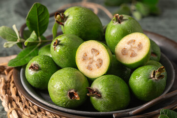 A close-up, moody still life of a metal bowl filled with green feijoa, with one fruit sliced in half, on a rustic wooden table