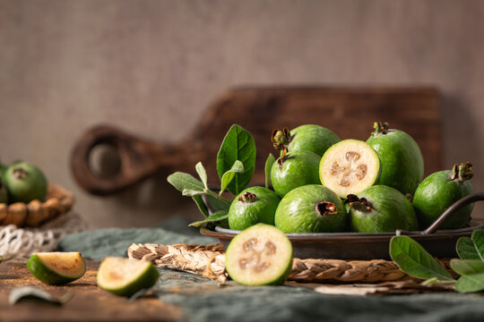 A fresh and rustic tabletop scene featuring feijoa fruit, a half-cut slice revealing seeds and pulp, presented in a metal plate.