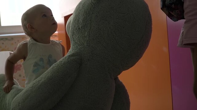 A baby girl with short blond hair in a blue suit lifts a huge blue teddy bear in her arms and looks at it with delight, smiles, and watches as her mother takes it and holds it in the air