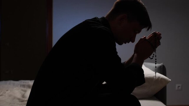 Young priest praying with rosary in dark room